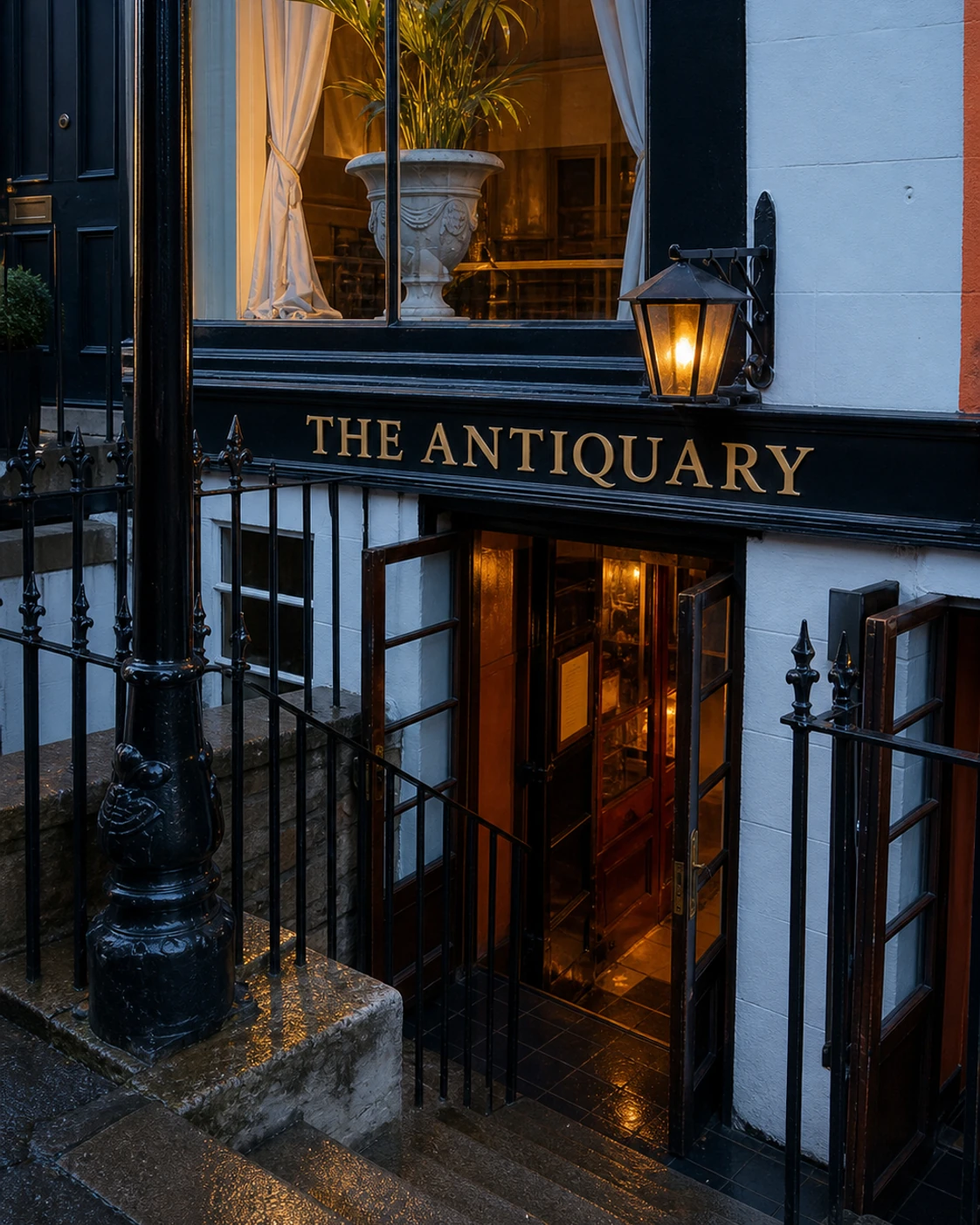 The Antiquary’s white and black basement entrance, iron railings and gold name above warmly lit steps.