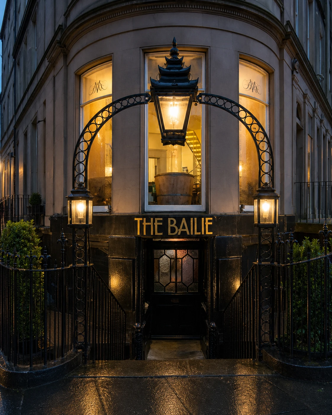 The Bailie’s black basement entrance beneath a curved iron arch, with the name in gold and warm lights reflected on wet paving.