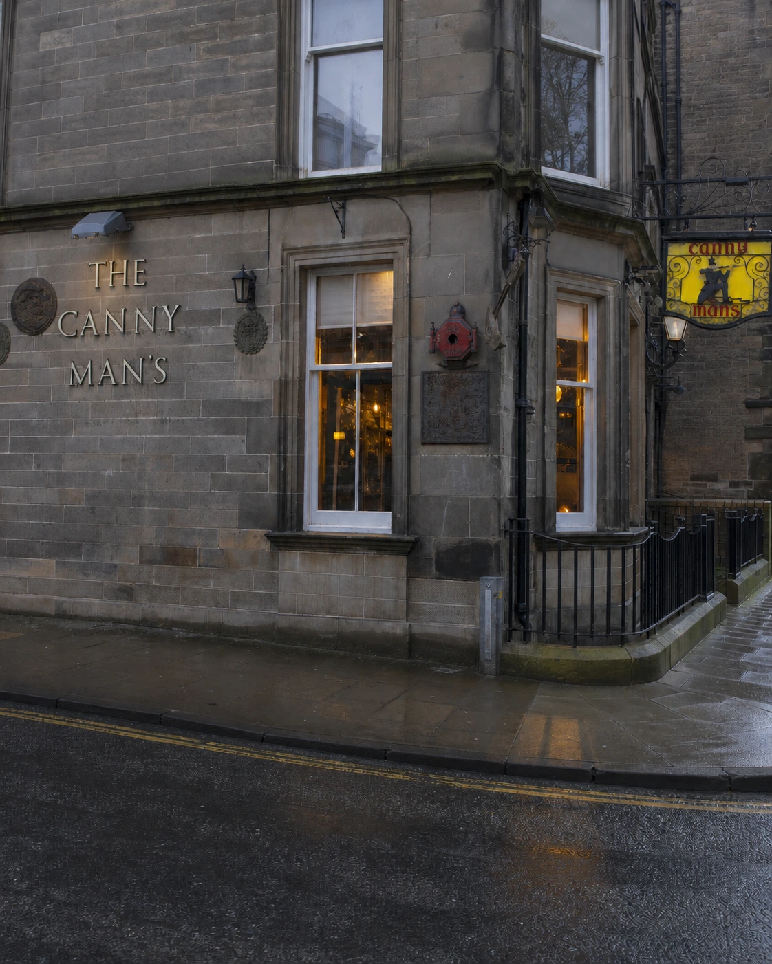 The Canny Man’s ashlar corner frontage with wall-mounted name letters, white sash windows and its yellow pictorial hanging sign after rain.
