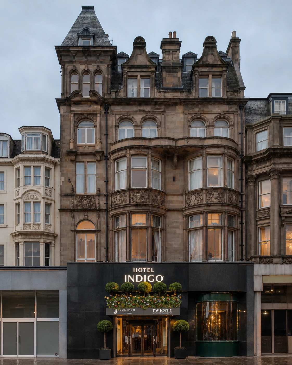 Juniper Edinburgh’s signed shared entrance beneath Hotel Indigo’s ornate sandstone 20 Princes Street façade, with topiary canopy and rain-darkened pavement.