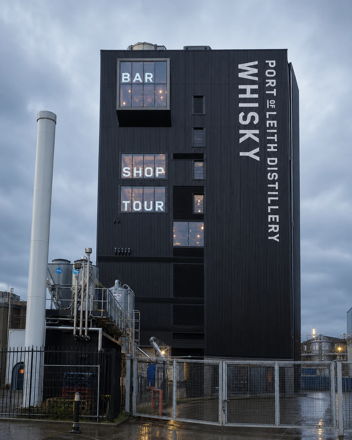 Port of Leith Distillery Bar’s exact black nine-storey waterfront tower, with BAR window, vertical white naming and rain-darkened industrial foreground.